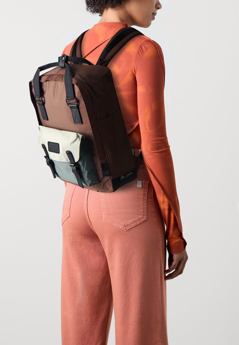 Hnědý batoh se černými popruhy, krémovou přední kapsou a spodní částí s zeleným akcentem. Nese jej osoba v oranžovém topu a růžových kalhotách.