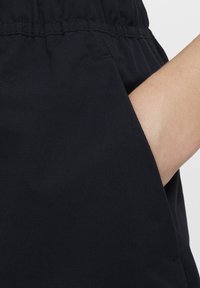 Črn material z gladko teksturo ima elastičen pas in stransko žep, kar poudarja funkcionalnost in minimalističen dizajn.