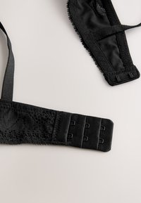 Schwarze Spitzen-BH-Träger mit Haken-und-Ösen-Verschlüssen flach auf einer hellen Oberfläche liegend, zeigt detaillierten Stoff und Nähte.