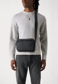 Schwarze Umhängetasche aus strapazierfähigem Material, mit einem Reißverschlussfach und verstellbarem Gurt, getragen über einem hellgrauen Pullover und dunklen Jeans.