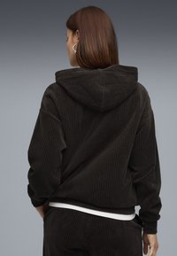 Felpa a righe in velluto a coste marrone con cappuccio e vestibilità rilassata, caratterizzata da una texture verticale a costine e un cinturino bianco che spunta sotto.