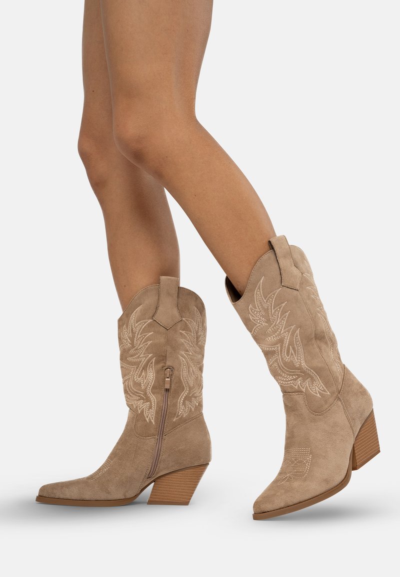 Botas de cowboy de ante beige con punteras puntiagudas, patrones florales bordados, altura hasta el tobillo y un tacón apilado. Cremallera lateral para facilitar su uso.