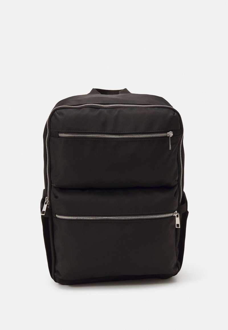 Sac à dos noir au design épuré, doté de deux poches avant zippées, d'un corps rembourré et d'accents en métal argenté. Texture lisse.