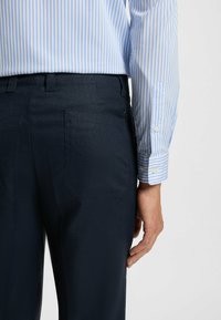 Marineblaue Hose aus strukturiertem Stoff, mit einer Gesäßtasche und einem gut definierten Taillenbund. Kombiniert mit einem hellblauen gestreiften Hemd.