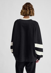 Person with short wavy hair wearing an oversized black sweatshirt with two white horizontal stripes on the sleeves, facing away.