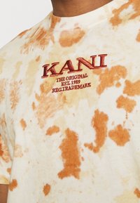 Detailní záběr na béžovou a oranžovou tričko s technikou tie-dye, na kterém je červený vyšitý text "KANI THE ORIGINAL EST. 1989 REG.TRADMARK" na hrudi.