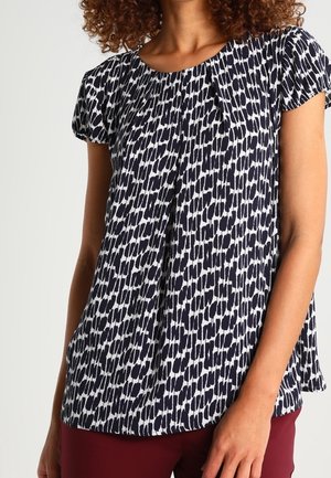 Kobieta ubrana w czarno-białą bluzkę w krótkim rękawie w wzory, do tego ciemnoczerwone spodnie, stojąca z jedną ręką spoczywającą wzdłuż ciała.