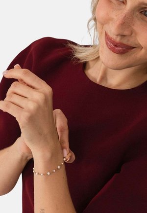 Woman with light skin and blonde hair wears a dark red top and adjusts a delicate gold bracelet with small pearls on her wrist.