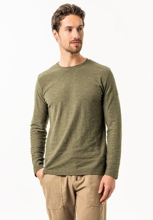 Man wearing olive green long-sleeve shirt and beige pants, standing with hands in pockets, looking to the side against a white background.