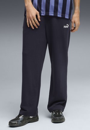 Person som bär svarta Puma mjukisbyxor, svarta och gråa sneakers och en blå och svart randig tröja, stående på grått golv.