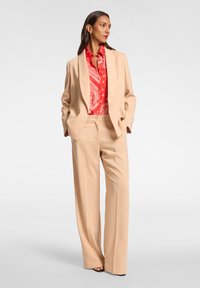 Blazer crema e pantaloni coordinati abbinati a una blusa rossa con motivo. Il blazer ha una vestibilità rilassata e i pantaloni sono a gamba larga.