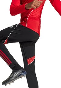 Sportoutfit bestehend aus einem roten Langarmshirt mit weißen Akzenten und schwarzen Hosen mit roten und weißen Details. Schwarze Fußballschuhe mit Stollen.