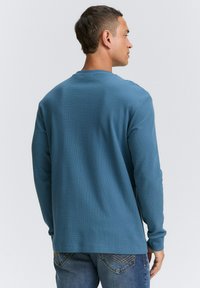 Camicia a maniche lunghe in tessuto blu testurizzato con scollatura rotonda, mostrata dalla retro e con maniche lunghe, abbinata a jeans denim.