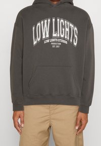 Una persona indossa un felpa grigia scura con la scritta "LOW LIGHTS" e ulteriori dettagli sul marchio, abbinata a pantaloni khaki.