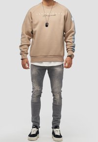 Beige sweatshirt med rund halsringning, logotyp på bröstet och blå ränder på ärmarna. Bärs över en vit skjorta, i kombination med slitna grå jeans och sneakers.