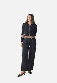 Zwart cropped lange mouwen top met ritssluiting, gedragen met zwarte wijde broek en schoenen met puntige neuzen. Gladde stof, minimalistisch ontwerp.
