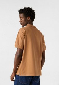 Polo marrone a maniche corte con colletto, spacchi laterali all'orlo e tessuto liscio e leggero. La vista posteriore mostra una vestibilità rilassata e cuciture.