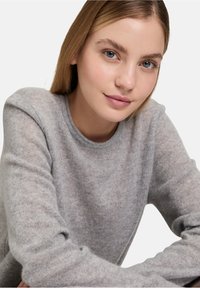 Szürke kötött pulóver kerek nyakkal és hosszú ujjakkal, puha textúrával és finom melírozott mintázattal. Közeli felvétel egy modell arcáról.