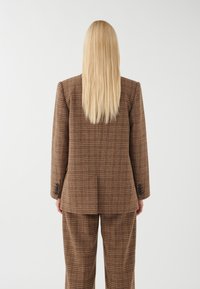 Brunternet blazer og bukser sæt, der har et struktureret stof, strukturerede skuldre og sorte knapdetaljer på ærmerne.