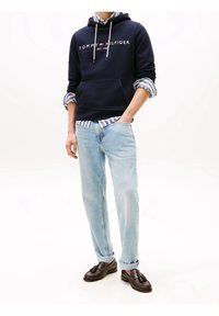 Felpa con cappuccio blu navy con logo bianco, jeans in denim azzurro chiaro e mocassini marroni. Indossata sopra una camicia a righe blu e bianche.