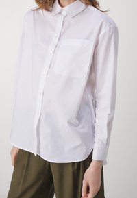 Donna che indossa una camicia bianca con bottoni e una tasca sul petto, insieme a pantaloni verde oliva, in piedi contro uno sfondo chiaro e uniforme.
