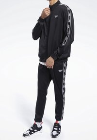 Černá sportovní souprava s mikinou na zip a zúženými kalhotami, obojí s bílými pruhy po stranách. Texturovaná látka a přiléhavý střih.