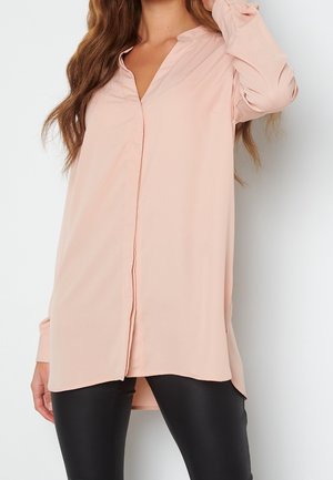 Mujer con una blusa suelta de color rosa pálido con botones y mangas largas, y pantalones ajustados negros, sobre un fondo liso.