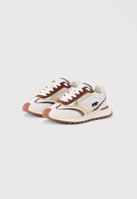 Witte en bruine veter-sneakers met rubberen zolen, metallic gouden accenten en een kleine afbeelding van een persoon met een zonnebril aan de zijkant.