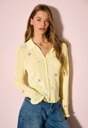 Keltainen kirjailtu pusero, jossa napillinen etuosa, pieniä kukka- ja sydänkuvioita sekä teksturoitu neulottu malli. Päällä sinisen farkun kanssa.