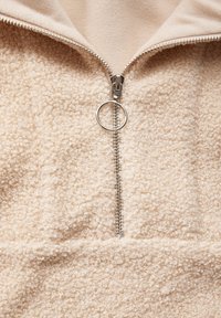 Felpa beige in pile con mezza zip e un tirante in metallo argentato. Il tessuto presenta un aspetto testurizzato e morbido, con una sensazione di calore.