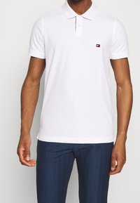 Bílá polo tričko s krátkým rukávem, dvouknoflíkovou légou a malým červeným a modrým logem na hrudi. Měkká, hladká tkanina a klasický design.