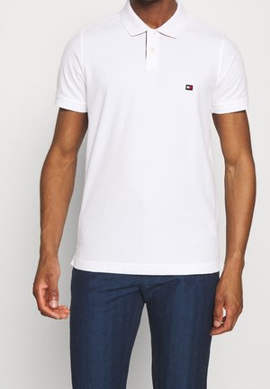 Polo shirt bianco con maniche corte, colletto a due bottoni e un piccolo logo rosso e blu sul petto. Tessuto morbido e liscio con un design classico.