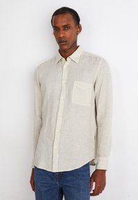 Camisa de lino beige claro de botones con un bolsillo en el pecho, mangas largas y dobladillo redondeado, combinada con jeans azules.