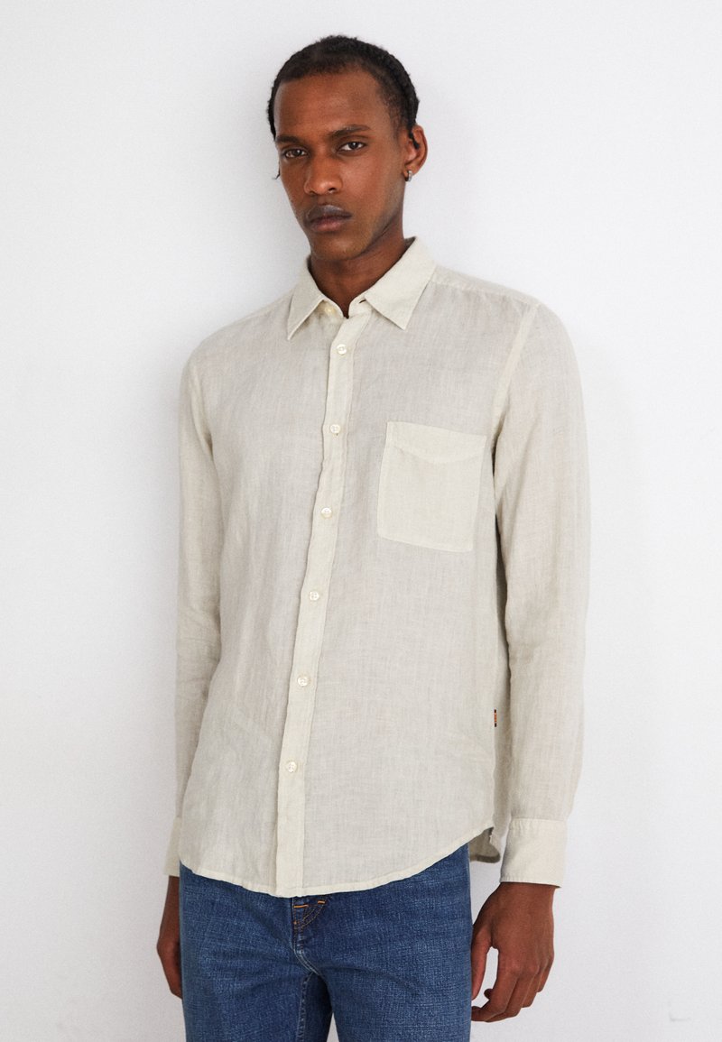 Camisa de lino beige claro de botones con un bolsillo en el pecho, mangas largas y dobladillo redondeado, combinada con jeans azules.