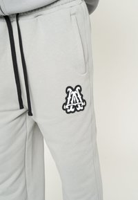 Sudadera gris con bolsillos laterales, cordones negros y un destacado logo bordado en blanco y negro en el muslo.