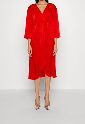 Mujer con un vestido rojo hasta la rodilla, de manga larga, escote en V y delantero drapeado, combinado con tacones altos plateados de punta.