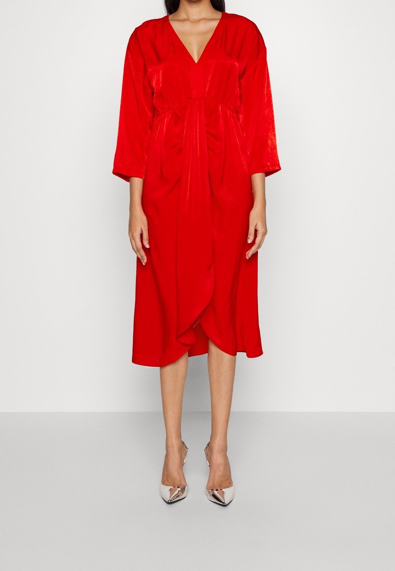 Femme portant une robe rouge mi-longue à manches longues, col en V et devant drapé, associée à des escarpins argentés à bout pointu.