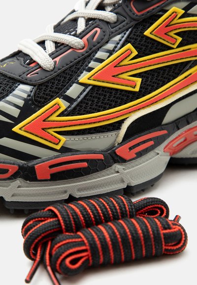 Μαύρο αθλητικό παπούτσι με κίτρινα και κόκκινα σχέδια βελών, λευκά κορδόνια και επιπλέον μαύρα και κόκκινα ριγωτά κορδόνια περασμένα δίπλα του.