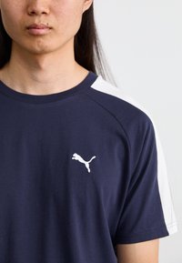 Marineblå t-shirt med kortærmer, hvid skulderdetalje og et lille hvidt logo på brystet. Stoffet virker let og glat.