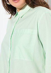 Camicia a righe verde chiaro con taschino sul petto, presenta un colletto strutturato e un bordo a contrasto lungo la pattina dei bottoni.