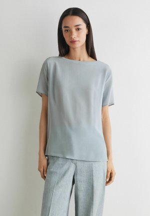 Ung kvinde med langt mørkt hår iført en lys blå løs kortærmet top og matchende lysegrå bukser med vide ben, står op ad en hvid væg.