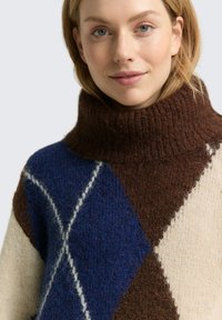 Kleurvolle gebreide trui met een hoge bruine kraag, versierd met grote blauwe en crèmekleurige diamanten patronen en een fuzzy textuur. Aangevuld met witte lijnen.