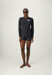 Haut noir à manches longues avec fermeture éclair à l'avant et logo brodé, associé à un short sombre. Texture lisse, design épuré et silhouette ajustée.