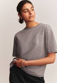 Camiseta de algodón gris con un bordado texturizado de un lazo plateado en la parte delantera, mangas cortas y un ajuste holgado.