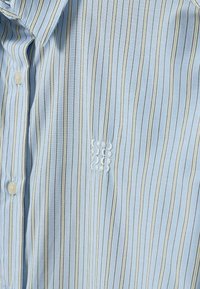 Chemise bleu clair à rayures verticales fines blanches, noires et jaunes, avec boutons sur le devant et logo blanc brodé sur la poitrine.