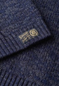 Gorro de punto en color azul marino con un patrón texturizado y una etiqueta dorada de Superdry. La vista en primer plano resalta los detalles del material y el tejido.