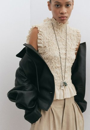 Joven mujer lleva blusa sin mangas color crema con volantes, falda plisada beige, chaqueta de cuero negra caída de los hombros y collar con colgante.