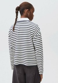 Henkilö, jolla on letitetyt hiukset, pukeutuneena valko-mustaraidalliseen pitkähiharaiseen paitaan ja tummaan housuun, selin yksinkertaista taustaa vasten.