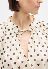 Γυναίκα που φοράει κρεμ μπλούζα με καφέ πουά, δέσιμο στον λαιμό και κοντά καστανά μαλλιά, στραμμένη προς τα δεξιά πάνω σε λευκό φόντο.