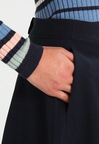 Jupe marine en tissu texturé, avec une poche latérale ; une main est placée à l'intérieur de la poche. La taille est lisse avec une fermeture discrète.